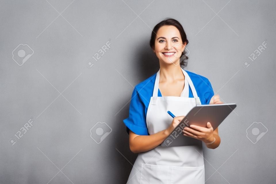 Happy smiling waitress taking orders isolated on grey wall. Mature woman wearing apron while writing on clipboard standing against gray background with copy space. Cheerful owner ready to take customer order while looking at camera. Small business concept.