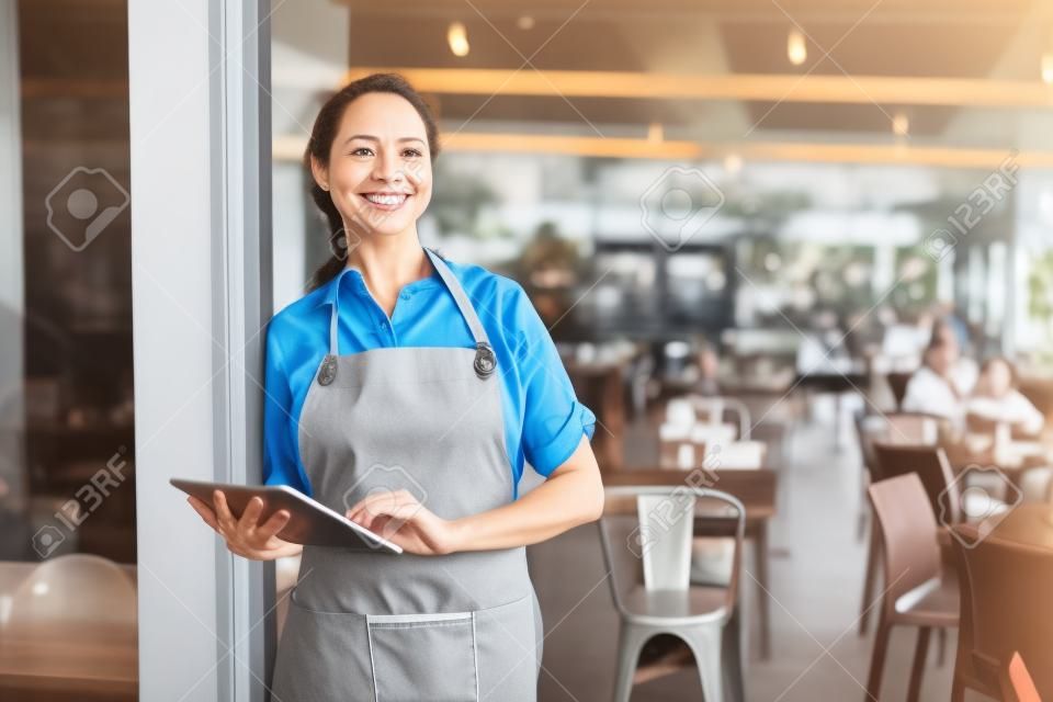 Portrait of a happy waitress standing at restaurant entrance holding digital tablet. Portrait of mature business woman standing at cafe entrance. Happy mature woman owner in grey apron standing at coffee shop entrance leaning while looking away with copy space.