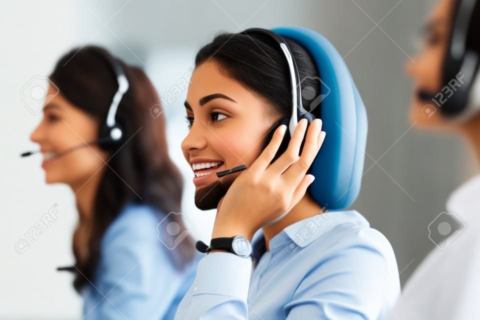 Multiethnic colleagues working in a call center. Team of business people sitting in a row working in a call center. Smiling customer support operator at work. Smiling young middle eastern woman with headset working at helpdesk.