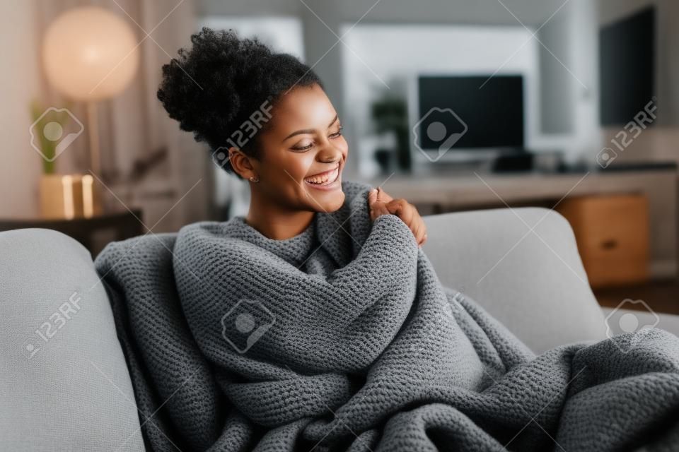 Bella ragazza nera seduta sul divano avvolta sotto una coperta e ridendo. Allegra donna afroamericana sensibile al freddo che si rilassa a casa sul divano. Metà donna spensierata e felice che si abbraccia con una coperta calda in inverno.