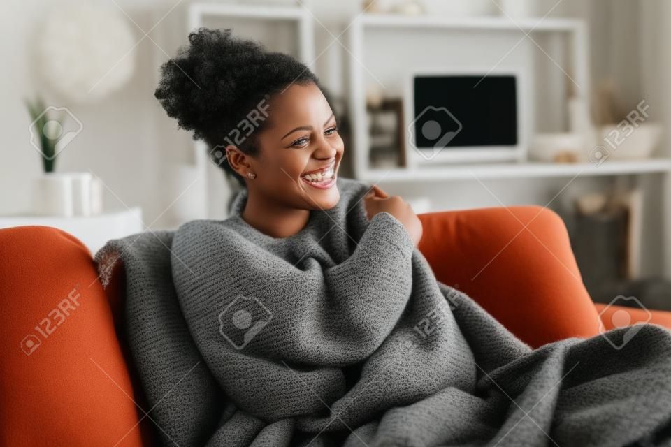 Schönes schwarzes Mädchen, das auf der Couch sitzt, unter Decke gehüllt und lacht. Fröhliche afroamerikanische Frau, die kälteempfindlich ist, entspannt sich zu Hause auf dem Sofa. Sorglose und glückliche mittlere Frau, die sich im Winter mit einer warmen Decke umarmt.