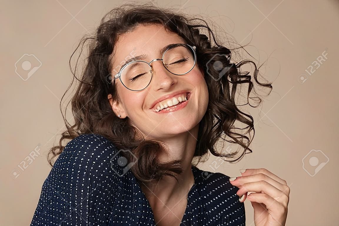 Casual cheerful woman with eyeglasses smiling at camera on cream background. Close up of happy young woman laughing with eyeglasses. Beautiful natural girl having fun with closed eyes showing a big grin.
