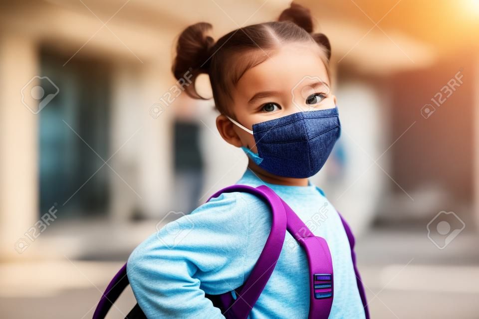 Smiling cute little girl with school backpack and protective face mask ready for first day of school during pandemic.