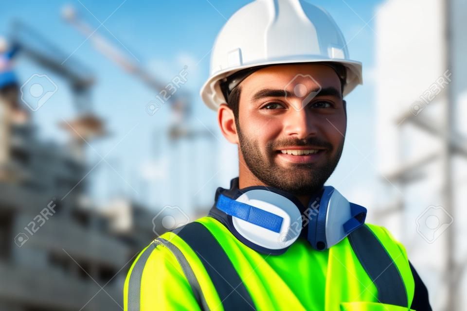 Porträt eines zufriedenen Baustellenleiters mit Sicherheitsweste und blauem Helm auf der Baustelle. Junger Architekt aus dem Nahen Osten beobachtet die Baustelle mit Zuversicht und blickt in die Kamera. Indischer Arbeiter auf einem Industriegebäude mit Kopienraum.
