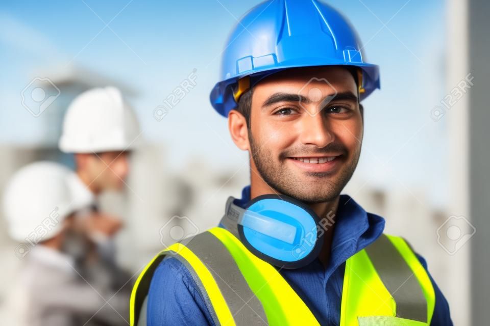 Porträt eines zufriedenen Baustellenleiters mit Sicherheitsweste und blauem Helm auf der Baustelle. Junger Architekt aus dem Nahen Osten beobachtet die Baustelle mit Zuversicht und blickt in die Kamera. Indischer Arbeiter auf einem Industriegebäude mit Kopienraum.