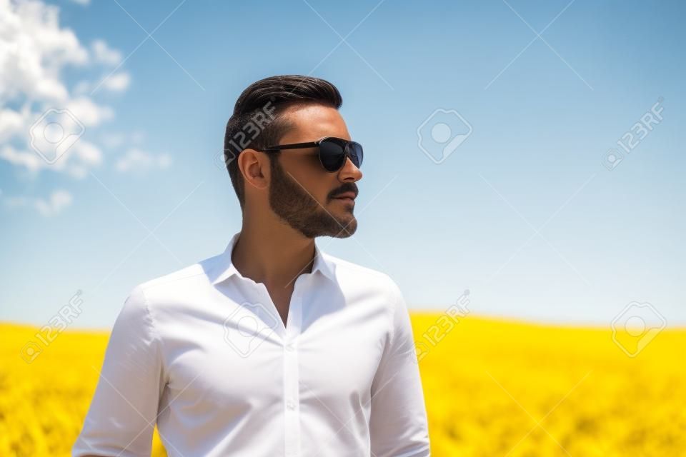 Um homem vestindo uma camisa branca e óculos escuros, em pé contra um grande campo amarelo olhando para longe da câmera. Um grande dia de verão ensolarado.