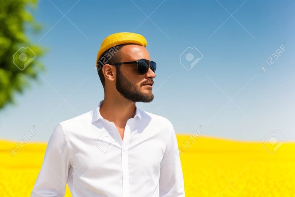 Un uomo che indossa una camicia scura e bianco occhiali da sole, in piedi contro un grande campo giallo guardando lontano dalla fotocamera. Una grande giornata di sole estivo.