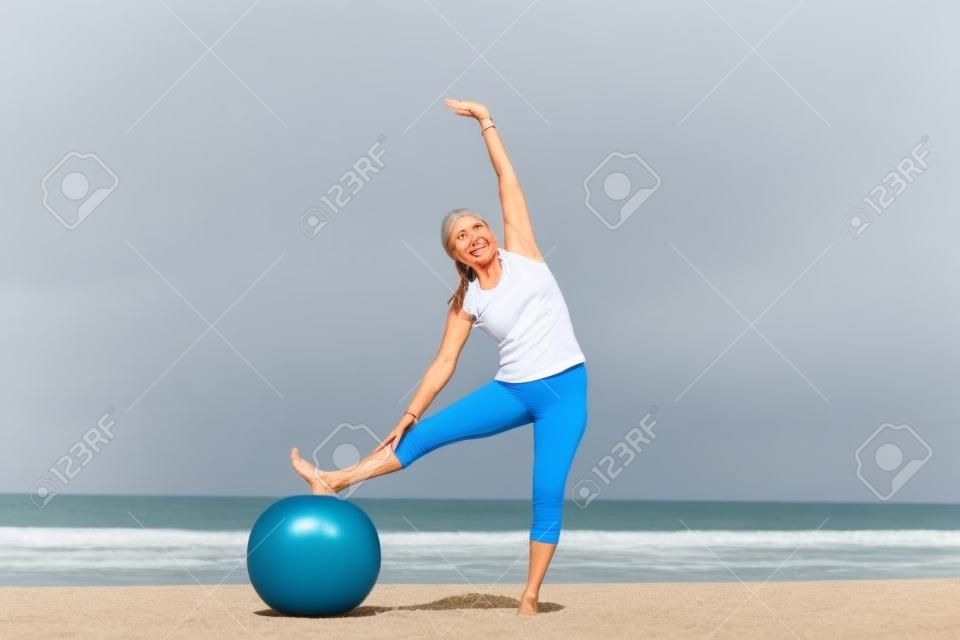 Fit and healthy attractive mature woman happy exercising with gymnastic ball at beach.