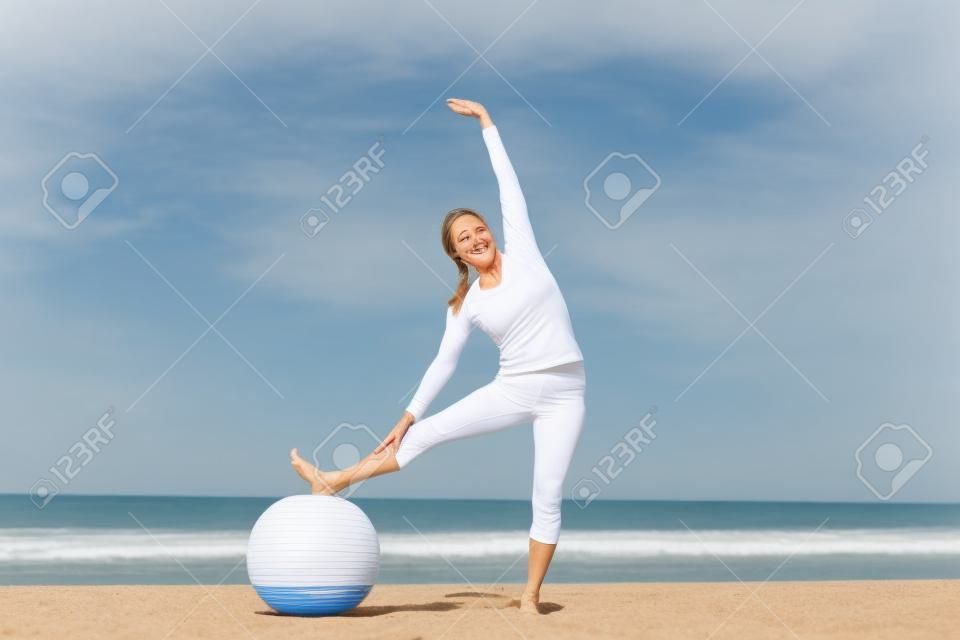 Здоровым привлекательным зрелая женщина счастлива упражнения с гимнастическим мячом на пляже.