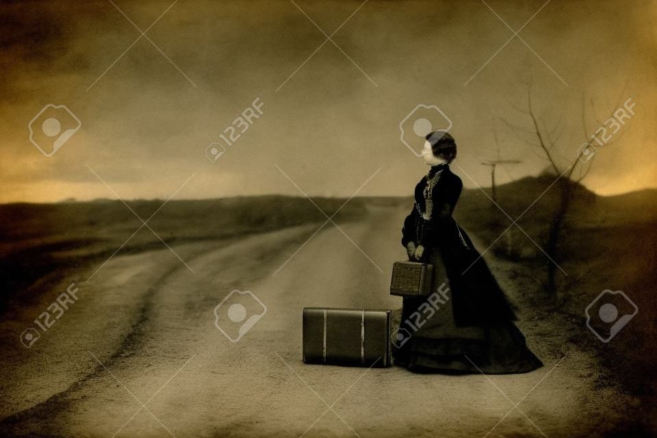 Retrato ao ar livre de uma senhora vitoriana de preto sentada sozinha na estrada com sua bagagem.