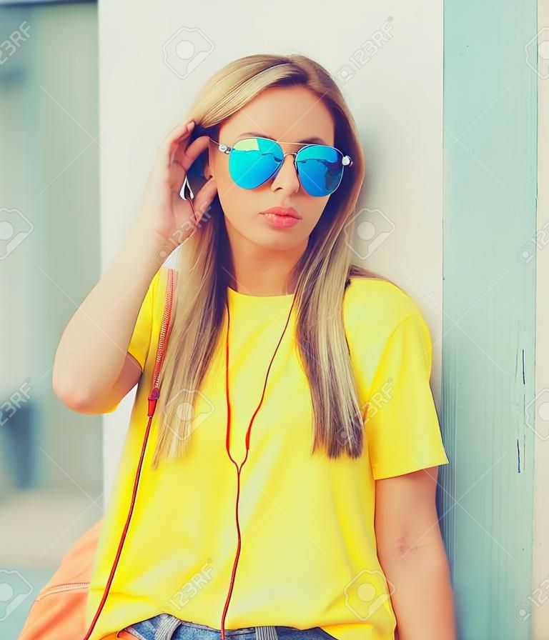 Porträt einer stilvollen, modernen blonden jungen Frau, die in der Stadt Musik über Kopfhörer hört und eine Sonnenbrille, einen Rucksack und ein gelbes T-Shirt trägt