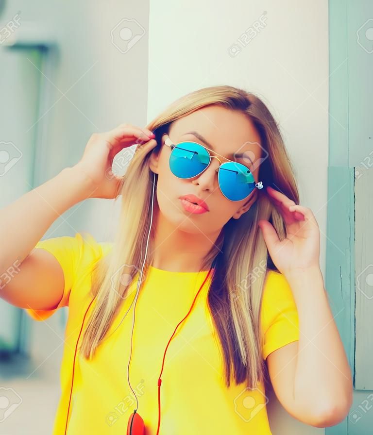 Portret van een stijlvolle, moderne blonde jonge vrouw die naar muziek luistert in een koptelefoon met een zonnebril, rugzak, geel t-shirt in de stad