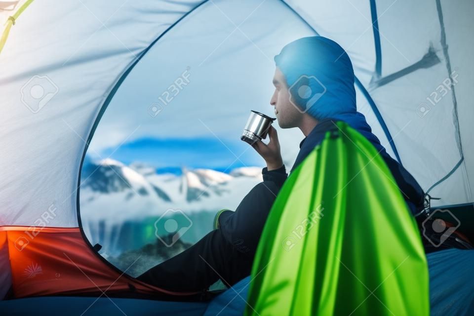 Il giovane escursionista beve il tè dalla boccetta in tenda al mattino