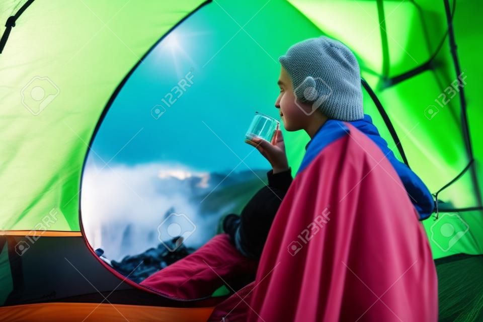 young hiker drinks tea from flask in tent in the morning