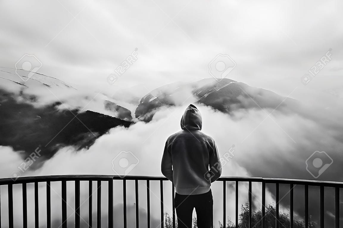 Widok z tyłu mężczyzny stojącego w punkcie widokowym, patrzącego na piękny krajobraz z mglistymi górami w oddali. Czarno-białe zdjęcie