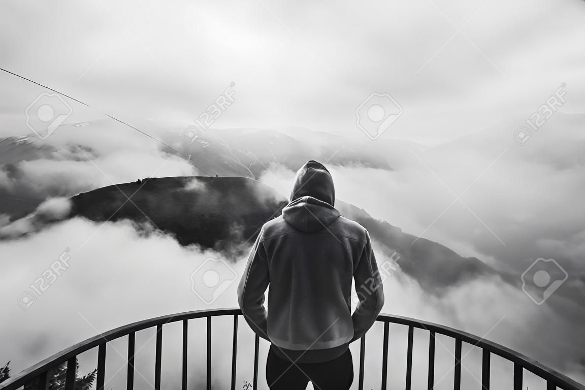 View from behind of a man standing at view point looking to beautiful landscape with foggy mountains in the distance. Black and white photo
