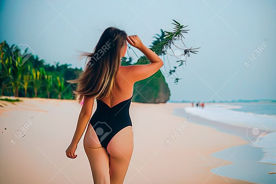 Vista posteriore del primo piano di una ragazza in costume da bagno nero che si gode il suo viaggio in spiaggia, godendosi il vento, il mare e la sabbia rosa, la vista dal retro. Il concetto di vacanze, vacanze, viaggi, persone