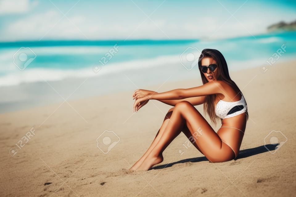 Sexy model in brown swimsuit and sunglasses posing on white sandy beach
