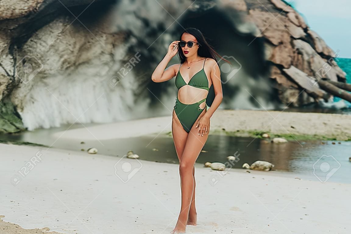 Schönes gebräuntes Mädchen in grüner Badebekleidung und Sonnenbrille posiert auf Strandhintergrund aus Felsen