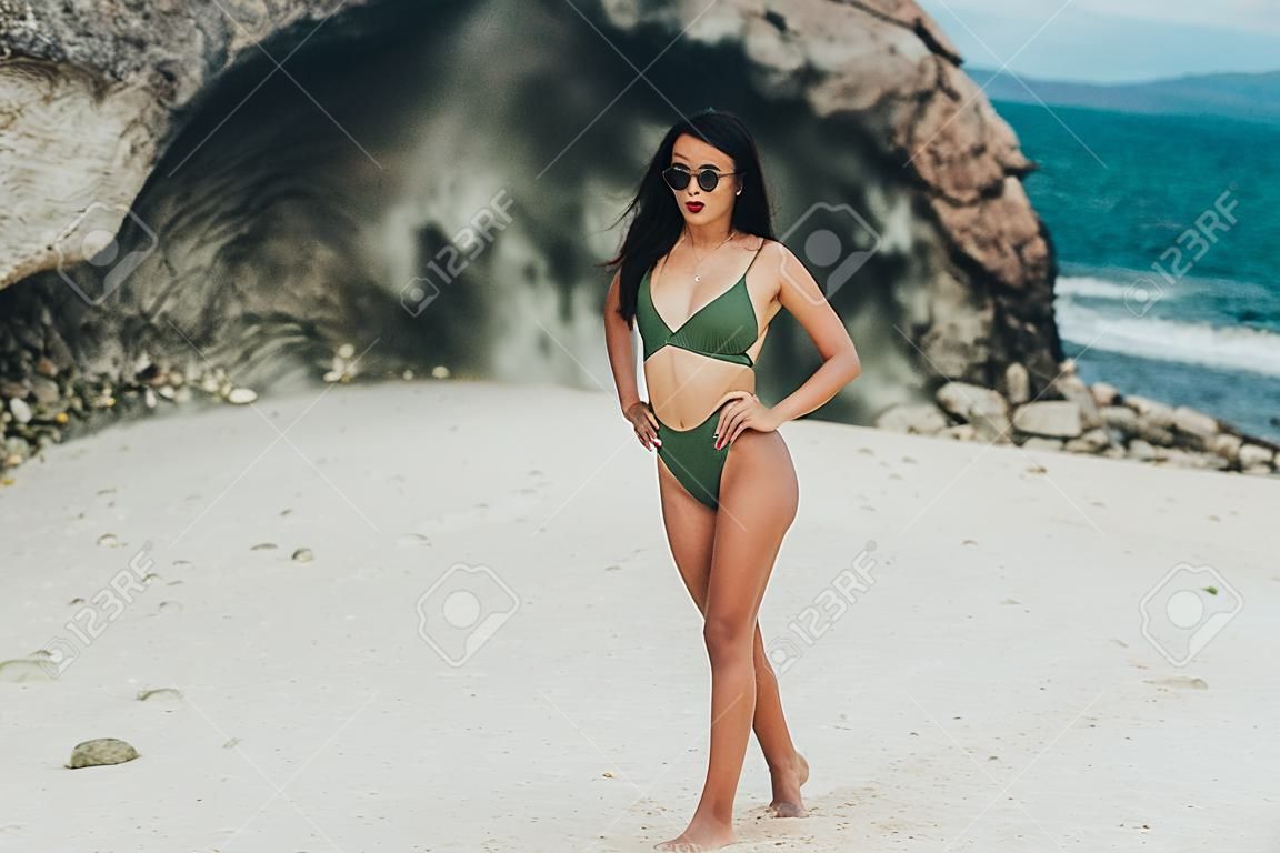 Beautiful tanned girl in green swimwear and sunglasses posing on beach background of rock