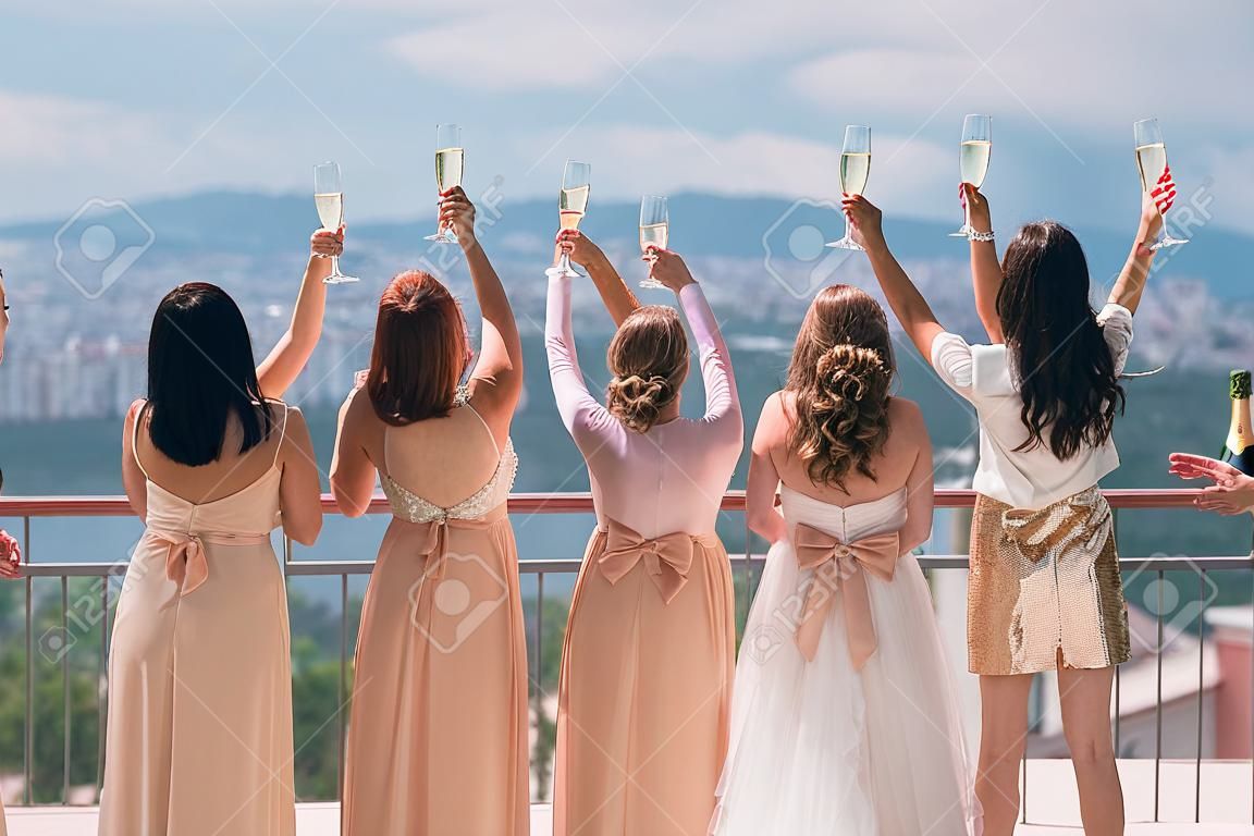 Vista posteriore sposa e fidanzate sul balcone, bicchiere di champagne sollevato