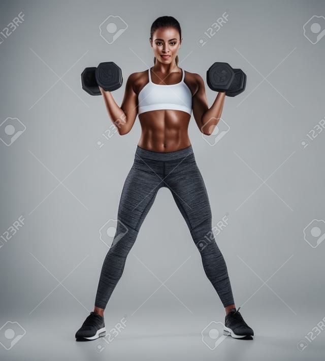 美麗的年輕女子在訓練中抽出背部肌肉和雙手啞鈴。照片運動的女人與完美的身體被隔絕在白色背景上。力量和動機