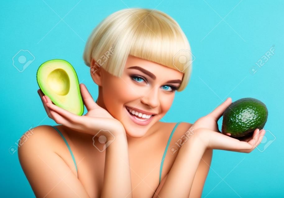 Happy girl with green avocado. Photo of smiling blonde girl on blue background. Skin care and beauty concept.