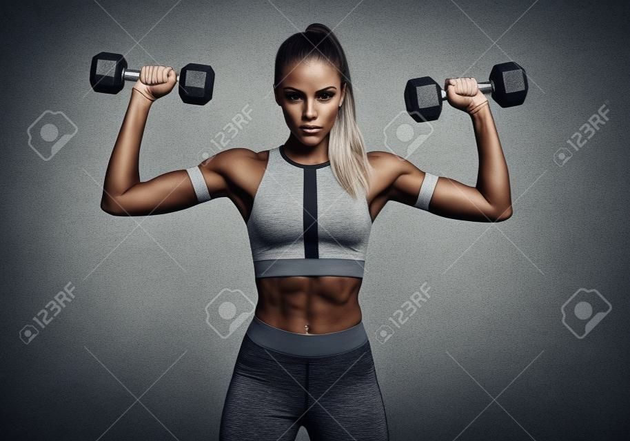 Hermosa joven haciendo ejercicios con pesas. Foto mujer atlética con cuerpo perfecto sobre fondo gris. Fuerza y motivación