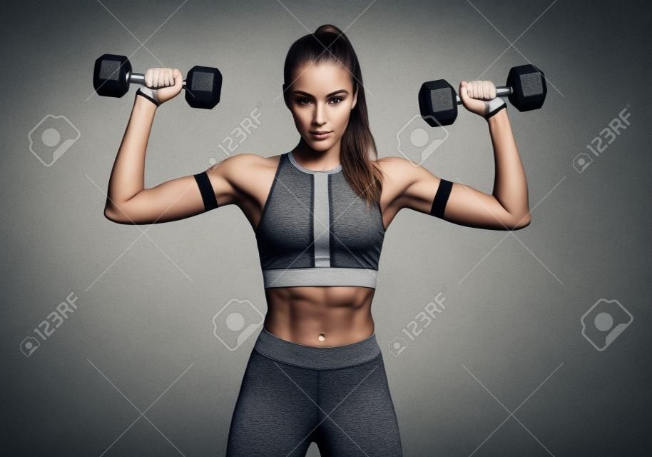 Hermosa joven haciendo ejercicios con pesas. Foto mujer atlética con cuerpo perfecto sobre fondo gris. Fuerza y motivación