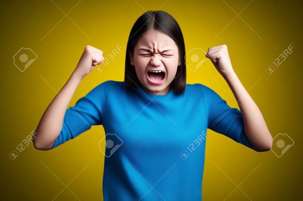 Aggression girl with widely open mouth and clenched teeth holds hands clenched in fists. Photo of girl in yellow sweater on blue background. Emotions and feelings concept