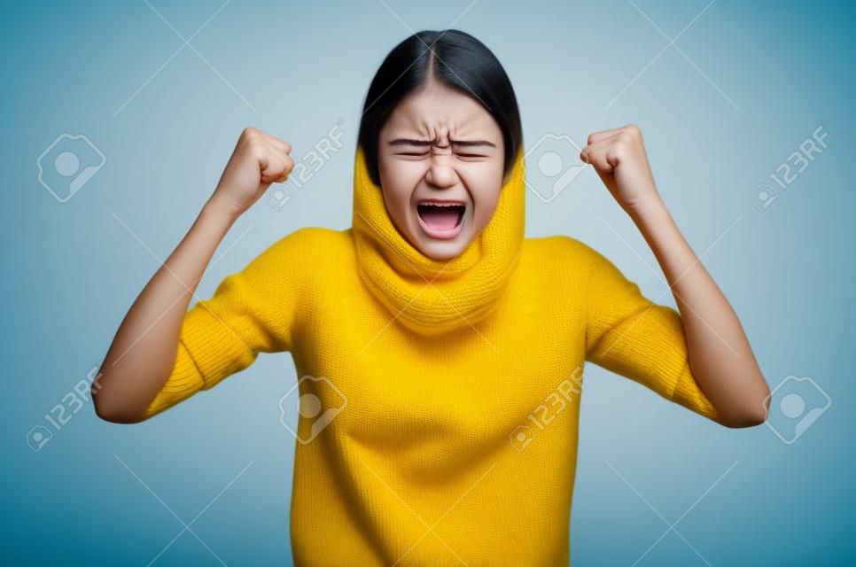 Aggression girl with widely open mouth and clenched teeth holds hands clenched in fists. Photo of girl in yellow sweater on blue background. Emotions and feelings concept