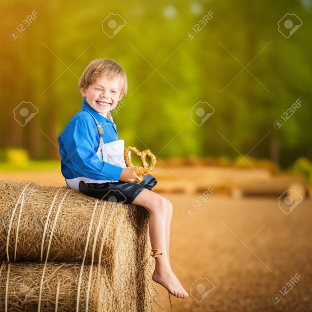 Lustiges kleines Kind Junge im traditionellen deutschen bayerischen Kleidung, Ledershorts und überprüfen Hemd, sitzen auf Heuhaufen oder Ballen und Essen Brezel. Aktiv im Freien Freizeit mit Kindern an einem warmen Sommertag.