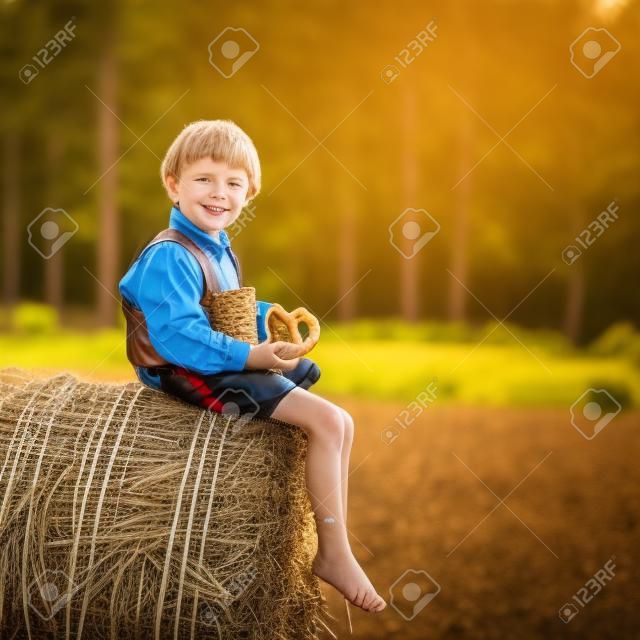 Lustiges kleines Kind Junge im traditionellen deutschen bayerischen Kleidung, Ledershorts und überprüfen Hemd, sitzen auf Heuhaufen oder Ballen und Essen Brezel. Aktiv im Freien Freizeit mit Kindern an einem warmen Sommertag.