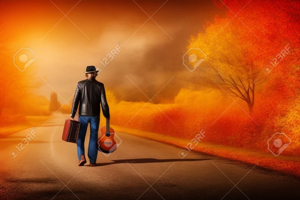 Молодой человек музыкант в стиле ретро шляпу и кожаной куртке с старинные чемодан и гитара уходить на пустые Осенняя дорога копией пространства фоне