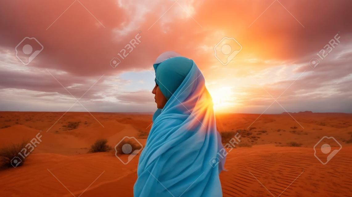Una giovane donna con il velo si trova nel deserto e guarda il tramonto