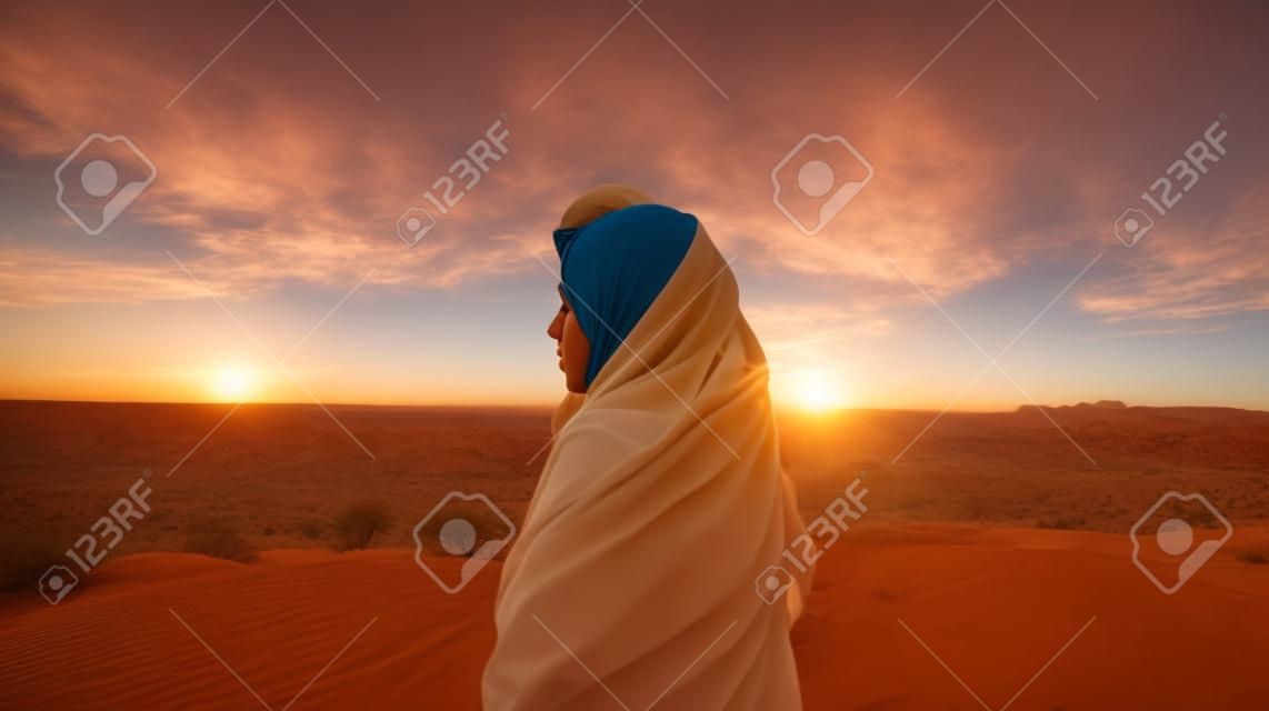Una mujer joven con un pañuelo en la cabeza se para en el desierto y mira la puesta de sol.