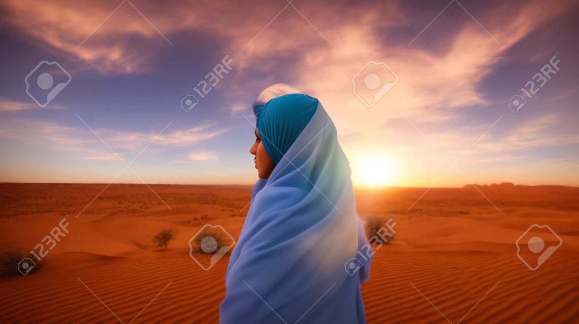A young woman in a headscarf stands in the desert and looks at the sunset.