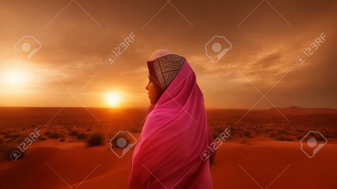 Een jonge vrouw met een hoofddoek staat in de woestijn en kijkt naar de zonsondergang