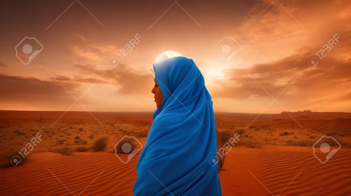Młoda kobieta w chustce stoi na pustyni i patrzy na zachód słońca