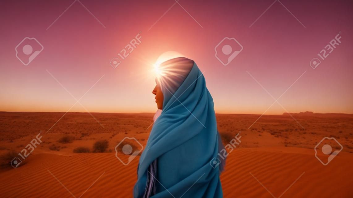 A young woman in a headscarf stands in the desert and looks at the sunset.