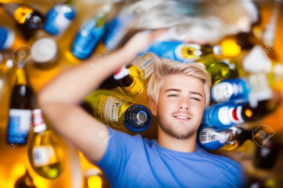 Young man with short blond hair lying on the floor and is surrounded by many empty beer and liquor bottles, upper perspective.