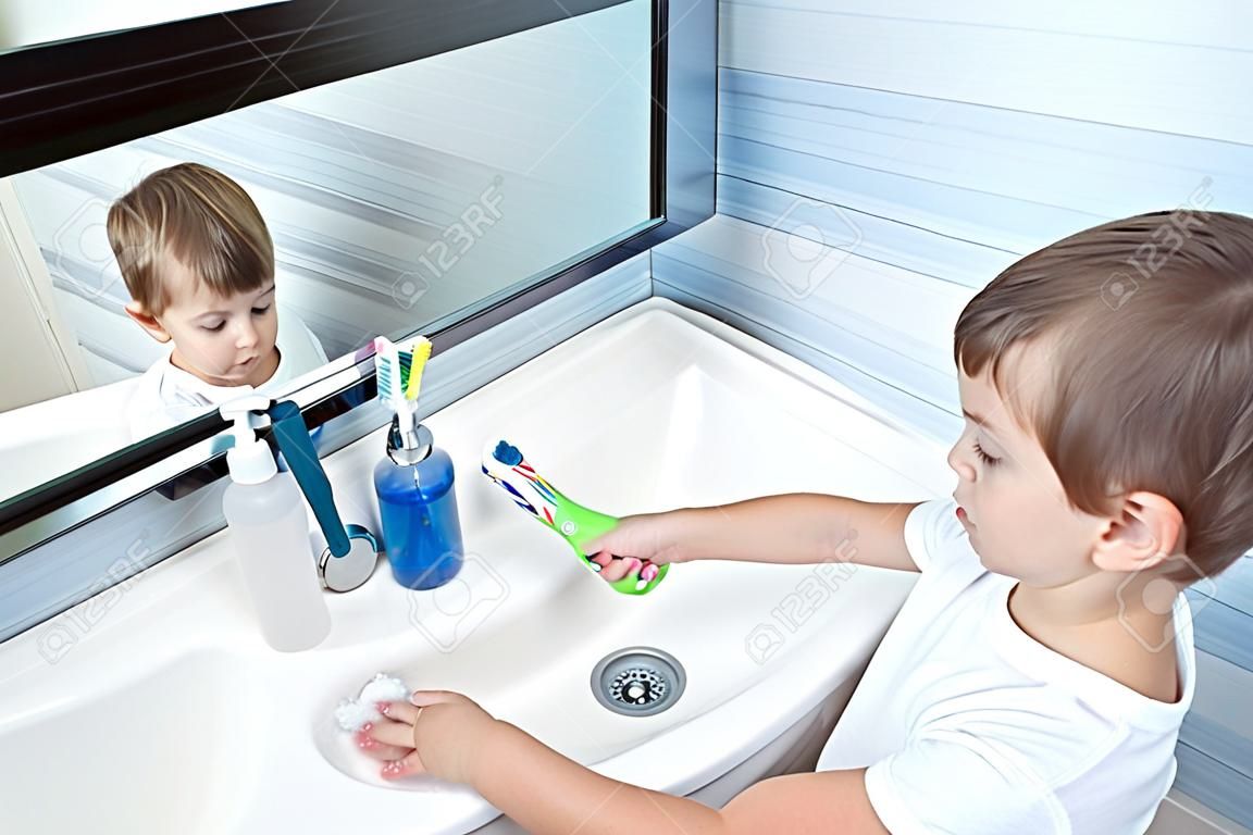 niño pequeño lindo en camiseta blanca que limpia su cepillo de sol bajo la corriente de agua en el baño