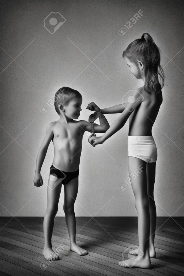 szczupła, zdrowa dziewczynka kaukaski bada silne mięśnie małego chłopca z miarką czarno-białe zdjęcie