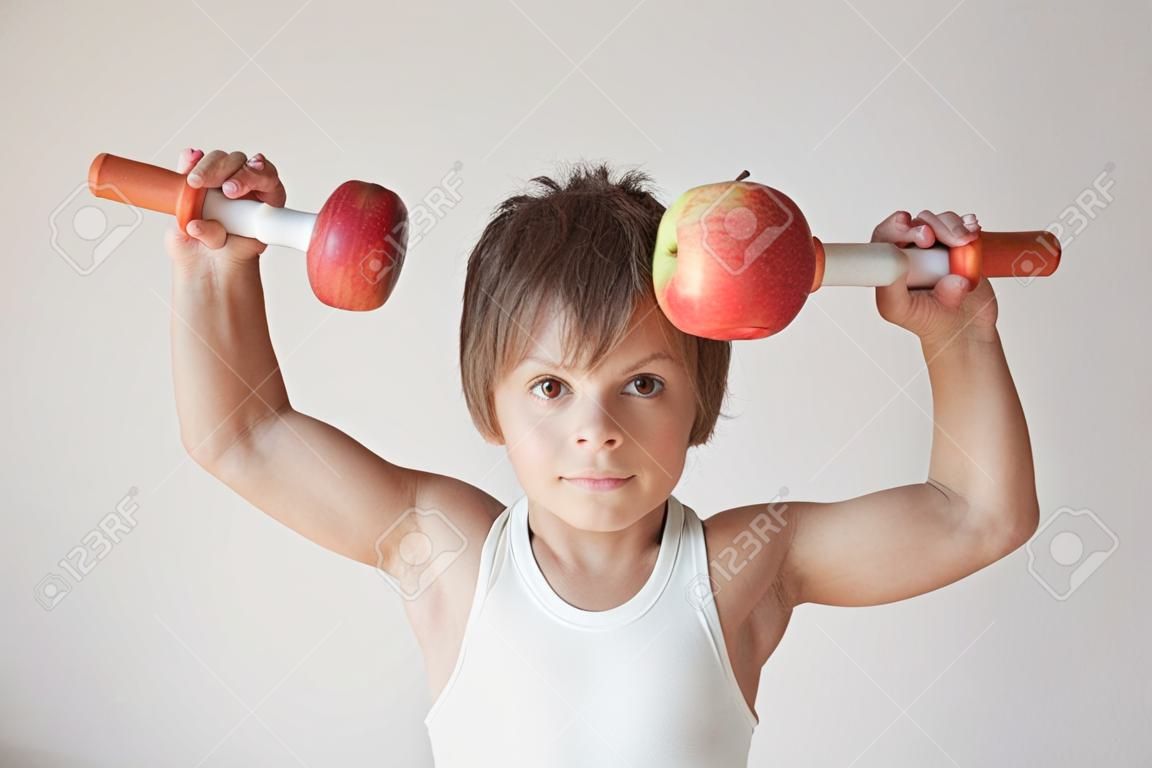 Süßer kleiner Junge im weißen Tanktop, der während des Sporttrainings Hanteln aus frischen Äpfeln hebt