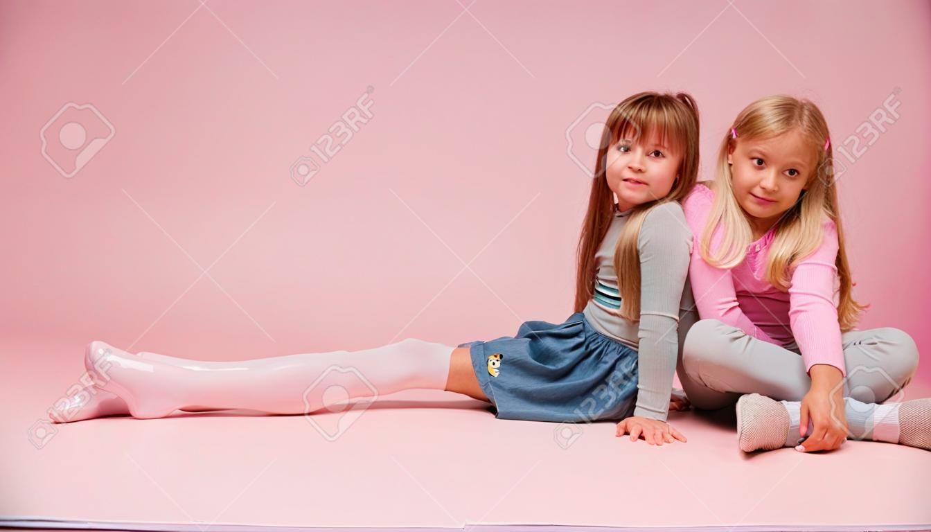 Due bambine carine sono sedute una accanto all'altra su uno sfondo rosa in studio. Scuola materna, infanzia, divertimento, concetto di famiglia. Una posa alla moda di due sorelle.