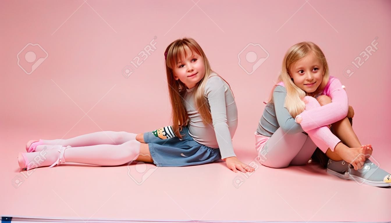 Two cute little girls are sitting next to each other on a pink background in the studio. Kindergarten, childhood, fun, family concept. Two fashionable sisters posing.