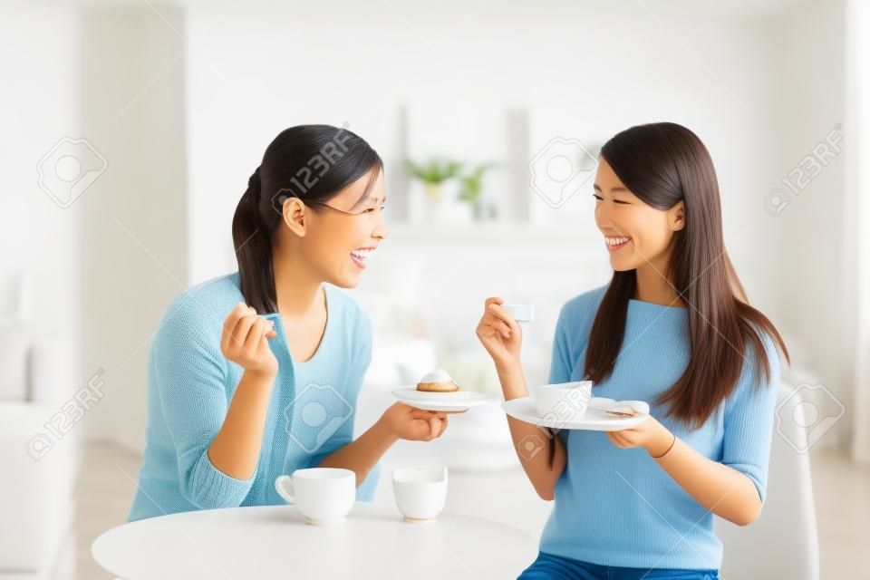 Два счастливых молодых женщин друзей с чашки кофе и пирожные, наслаждаясь разговор в гостиной дома, концепция здорового образа жизни, азиатской красоты