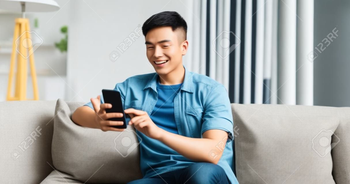Asiatischer junger mann, der auf dem sofa sitzt, überprüft die sozialen medien per handy glücklich im wohnzimmer zu hause