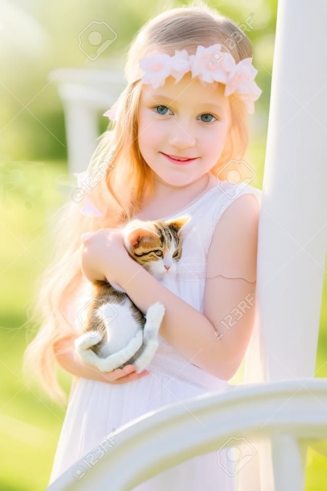 부드러움과 고양이를 사랑하고 행복으로 웃는 작은 귀여운 아이의 초상화. 그녀의 새끼 고양이와 함께 공원에서 편안한 어린 소녀. 하얀 친교 드레스를 입은 아이가 고양이에게 키스하고 있다. 봄 패션