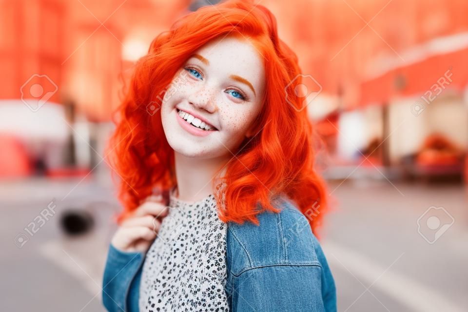 Portrety uroczej rudowłosej dziewczyny z piegami i ładną twarzą, którą dziewczyna pozuje do kamery w centrum miasta, ma świetny nastrój i słodki uśmiech